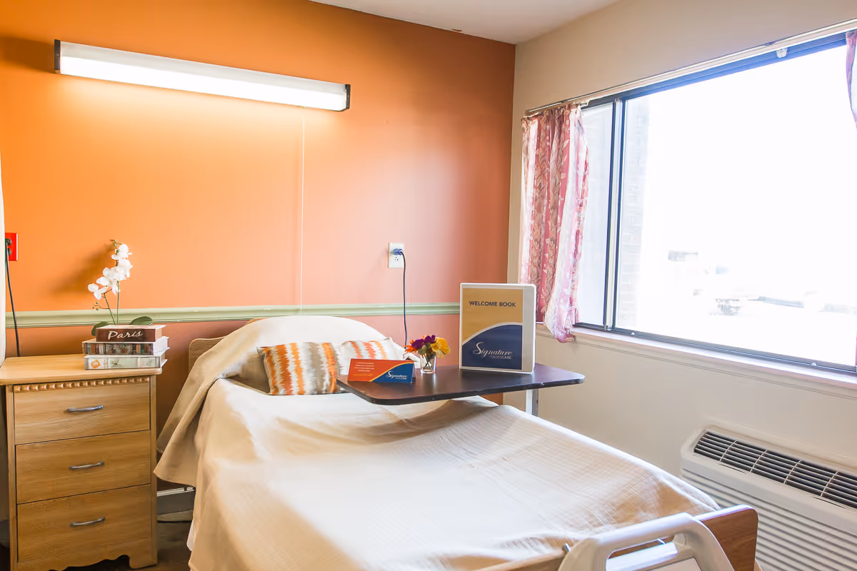 A single bed in a room with an orange accent wall and a large window with pink patterned curtains. The bed has a white blanket and a decorative pillow. Next to the bed is a wooden nightstand with a small stack of books and a white orchid plant. A small table is positioned over the bed holding a welcome book and a small vase with flowers. There is a wall-mounted light fixture above the bed and an air conditioning unit below the window.