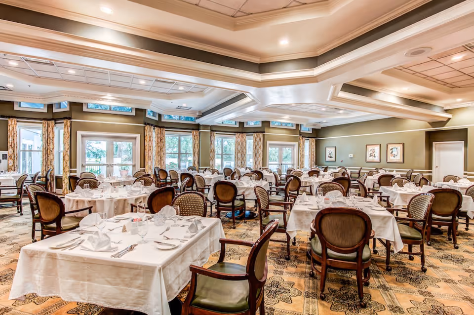 A spacious dining room with multiple round and square tables covered with white tablecloths, set with glassware, napkins, and silverware. The room features large windows with patterned curtains, green walls, decorative ceiling molding, and patterned carpet flooring. Wooden chairs with cushioned seats surround the tables.