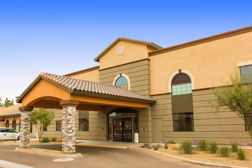 Exterior front entrance of a tan stucco building with a tiled covered porte-cochère, arched windows, and desert landscaping.