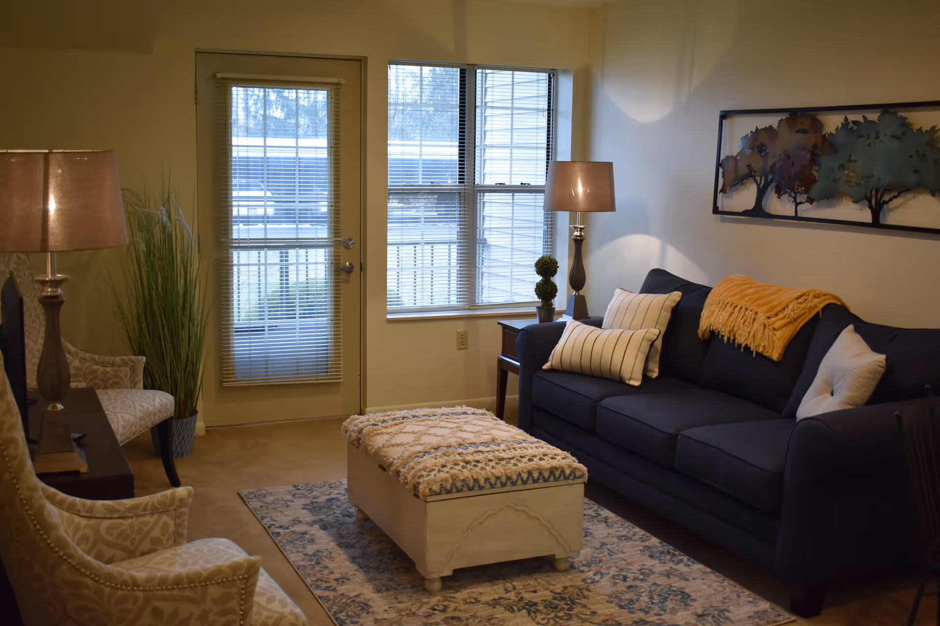 A cozy living room with a dark blue sofa adorned with striped and solid pillows and a yellow throw blanket. In front of the sofa is a cushioned ottoman on a patterned rug. Two beige patterned armchairs are positioned opposite the sofa. There are two table lamps on either side of the sofa, a small side table with decorative plants, and a large window and door with blinds letting in natural light. A framed artwork featuring trees hangs on the wall above the sofa.