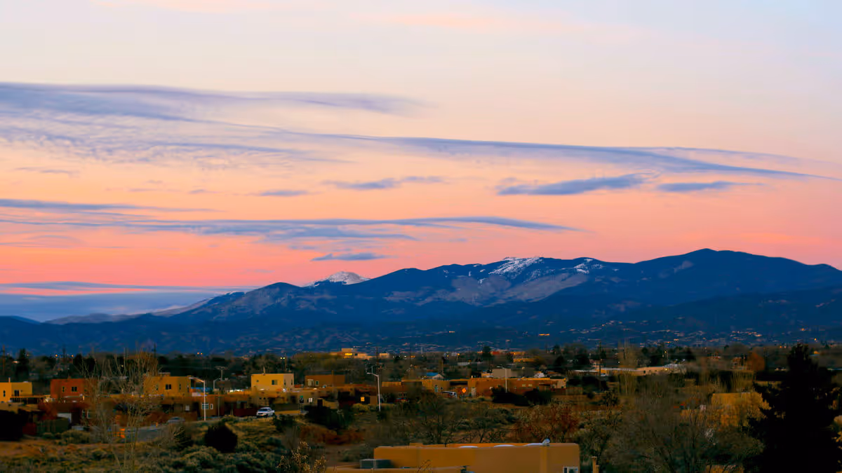 Scenic view of a residential area with southwestern-style buildings in the foreground and a mountain range with some snow patches in the background under a colorful sunset sky.