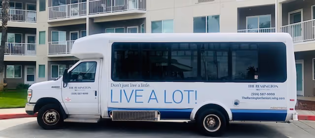 A white shuttle bus parked in front of a senior living facility building. The bus has the text 'Don't just live a little. LIVE A LOT!' in large blue letters on its side, along with the facility name 'THE REMINGTON', a phone number, and a website URL.