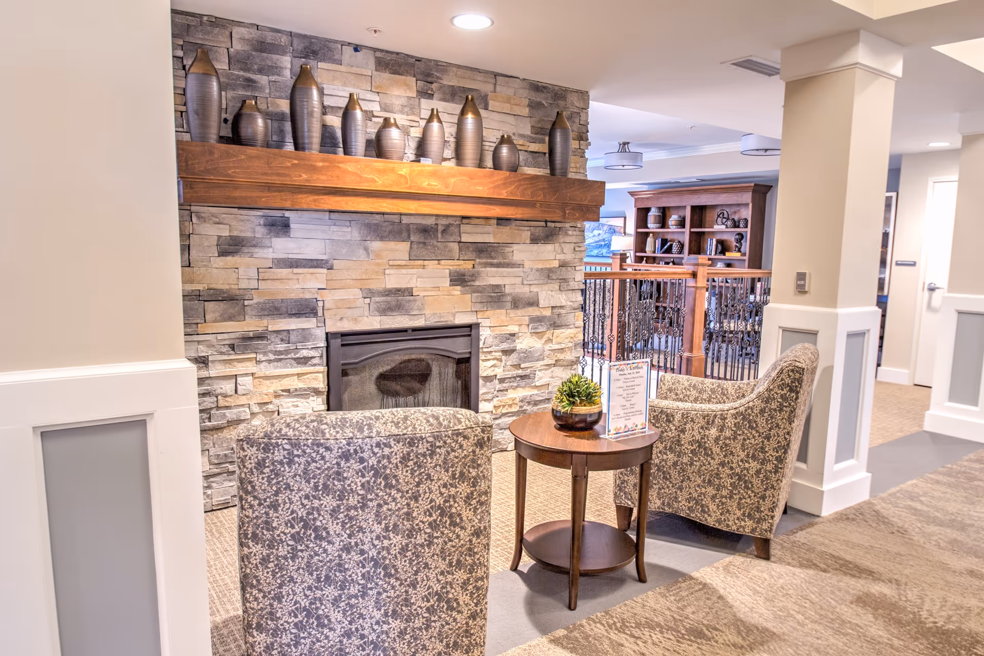A cozy seating area in a senior living facility featuring two patterned armchairs facing a stone fireplace with a wooden mantel decorated with vases. A small round wooden table between the chairs holds a small plant and a sign. In the background, there is a wooden bookshelf and a railing with decorative ironwork.