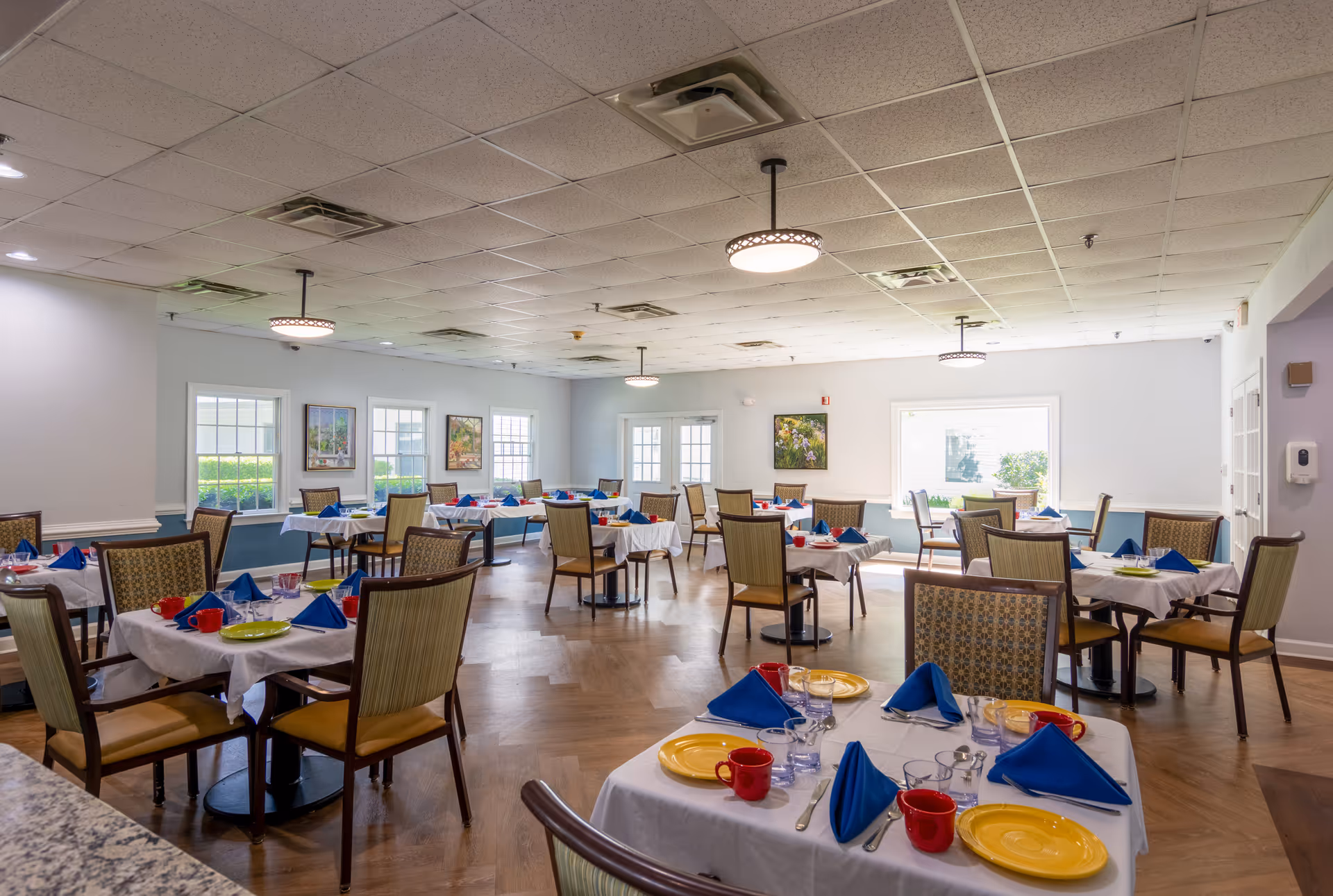 Bright communal dining room with multiple tables set with white tablecloths, blue napkins, colorful plates and cups, and chairs arranged for residents.