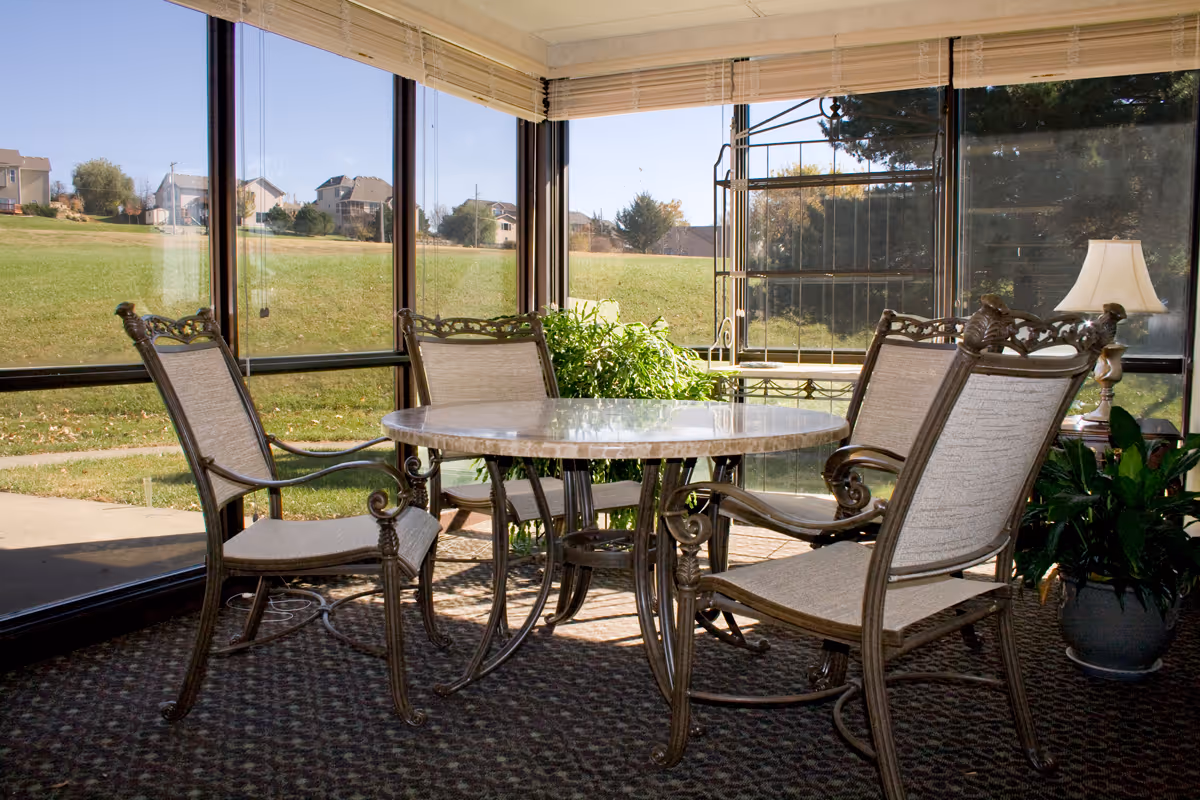 Sunlit enclosed patio/sunroom with a round table and four ornate metal-framed chairs facing large windows overlooking a grassy lawn.
