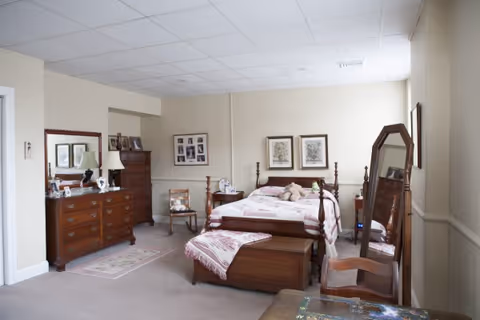 A bedroom with a wooden bed featuring a pink and white quilt and a teddy bear on top. There is a wooden dresser with a mirror, a chest of drawers, a wooden chair, and a full-length mirror. The walls are light-colored and decorated with framed pictures.