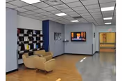 A spacious interior common area with light wood flooring and a white ceiling with fluorescent lights. There are two beige armchairs positioned near a wall with a bookshelf filled with books and decorative items. A flat-screen TV is mounted on a blue accent wall, displaying a news program. The hallway extends to the right with a door at the end.