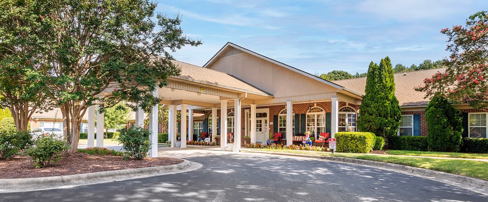 Front exterior view of a single-story senior living facility building with a covered entrance, surrounded by trees, shrubs, and a curved driveway under a partly cloudy sky.