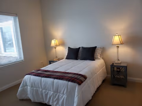 A neatly made bed with a white comforter, two black pillows, and a red plaid throw blanket at the foot. The bed is flanked by two bedside tables, each with a lit table lamp. There is a window with blinds on the left side of the room, and the walls are painted a light neutral color.