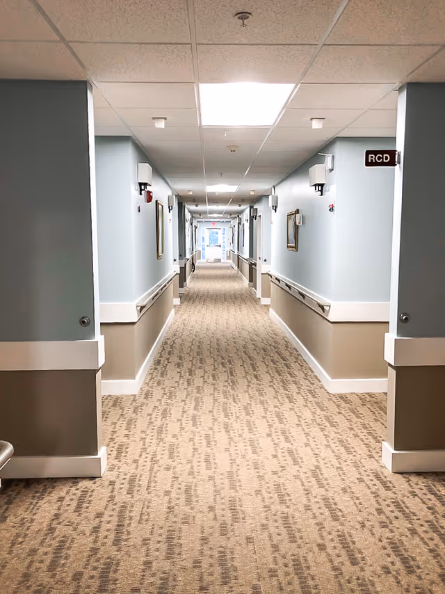 A long, well-lit hallway in a senior living facility with beige patterned carpet, light blue and beige walls, handrails on both sides, and framed pictures hanging on the walls. The ceiling has recessed lighting panels and smoke detectors. A sign labeled 'RCD' is visible on the right wall near the foreground.