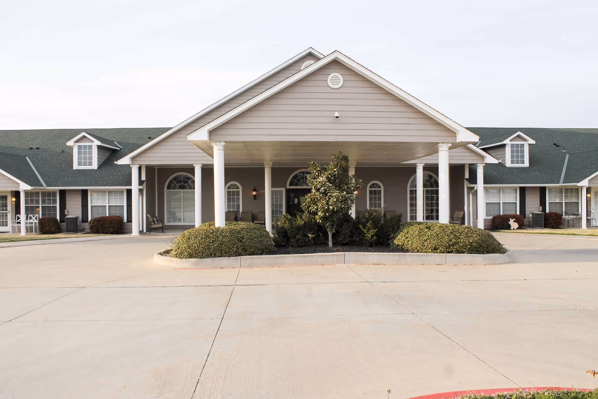 Front exterior view of a single-story building with a covered entrance supported by white columns, surrounded by bushes and a small tree in the center. The building has multiple windows with white frames and a dark green roof with dormer windows.