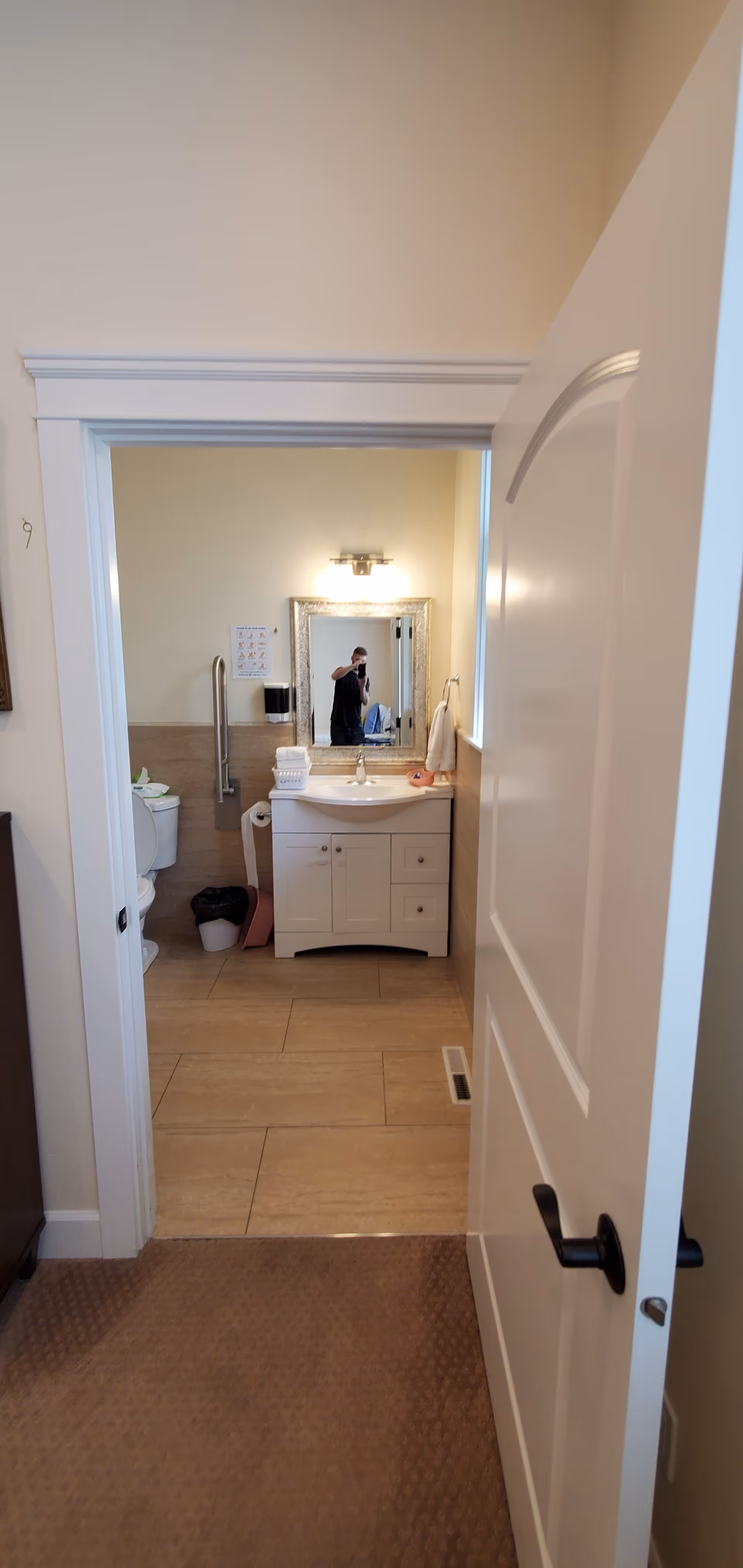 View through an open door into a bathroom with beige tiled floor and walls. The bathroom features a white vanity with a sink, a mirror above it, a light fixture, a toilet with a grab bar, and various bathroom accessories including a towel and a trash bin. A person is visible in the mirror taking the photo.