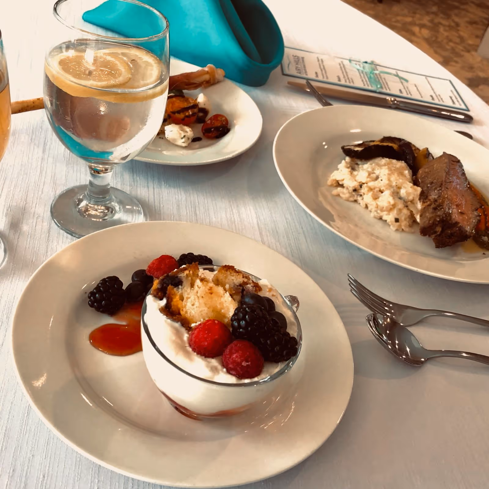 A table set with plates of food including a dessert with whipped cream and berries, a plate with meat and a creamy side dish, and a small plate with grilled vegetables and cheese. There is also a glass of water with a lemon slice and silverware on a white tablecloth.