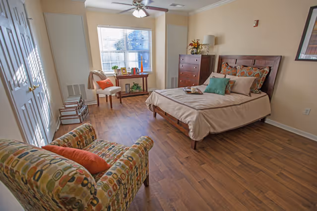 Sunlit furnished bedroom with a bed, patterned armchair, dresser, and a window with blinds.