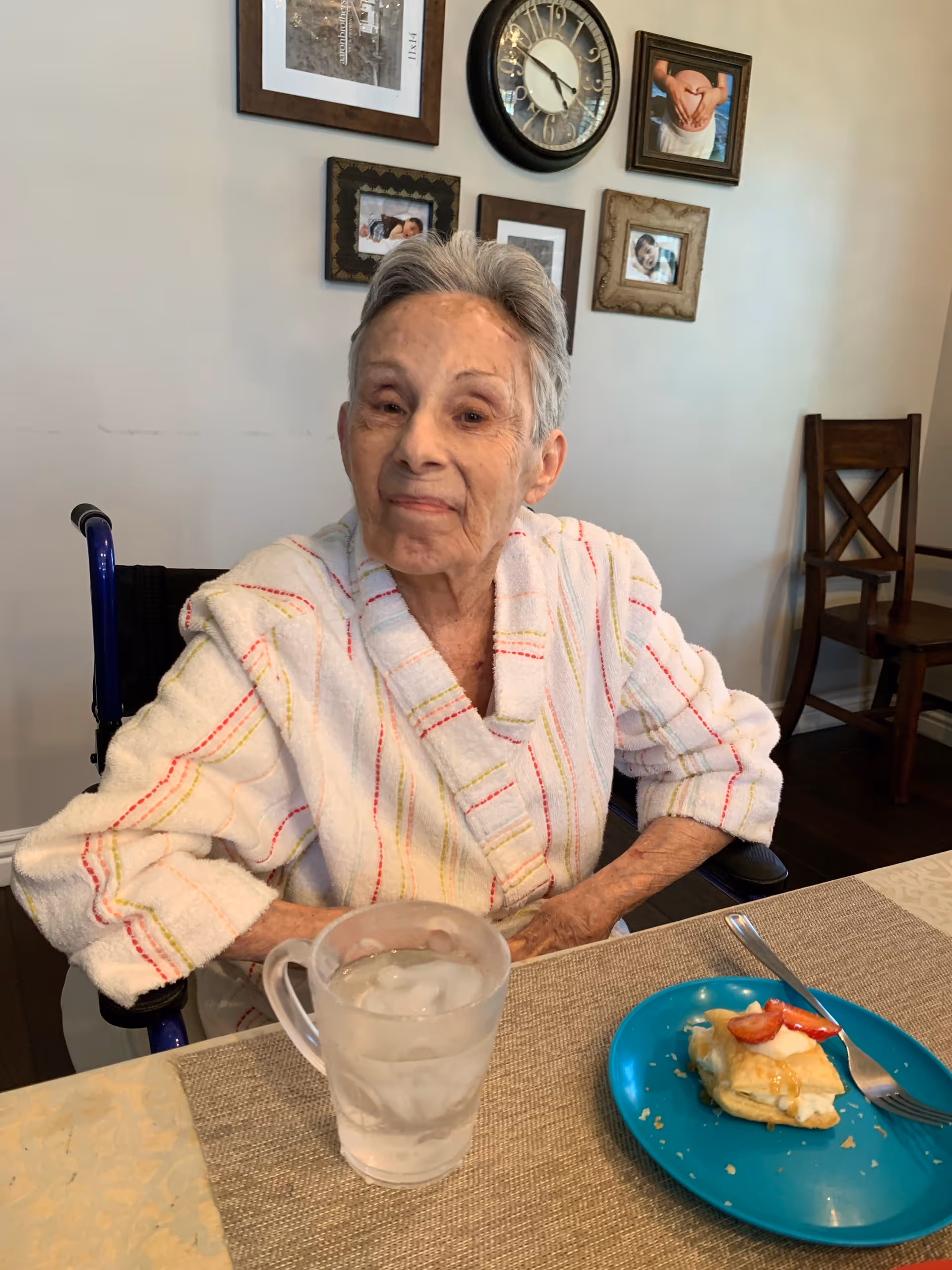 An elderly woman in a robe sits at a dining table with a glass of ice water and a plate with a pastry, with a clock and framed photos on the wall behind her.