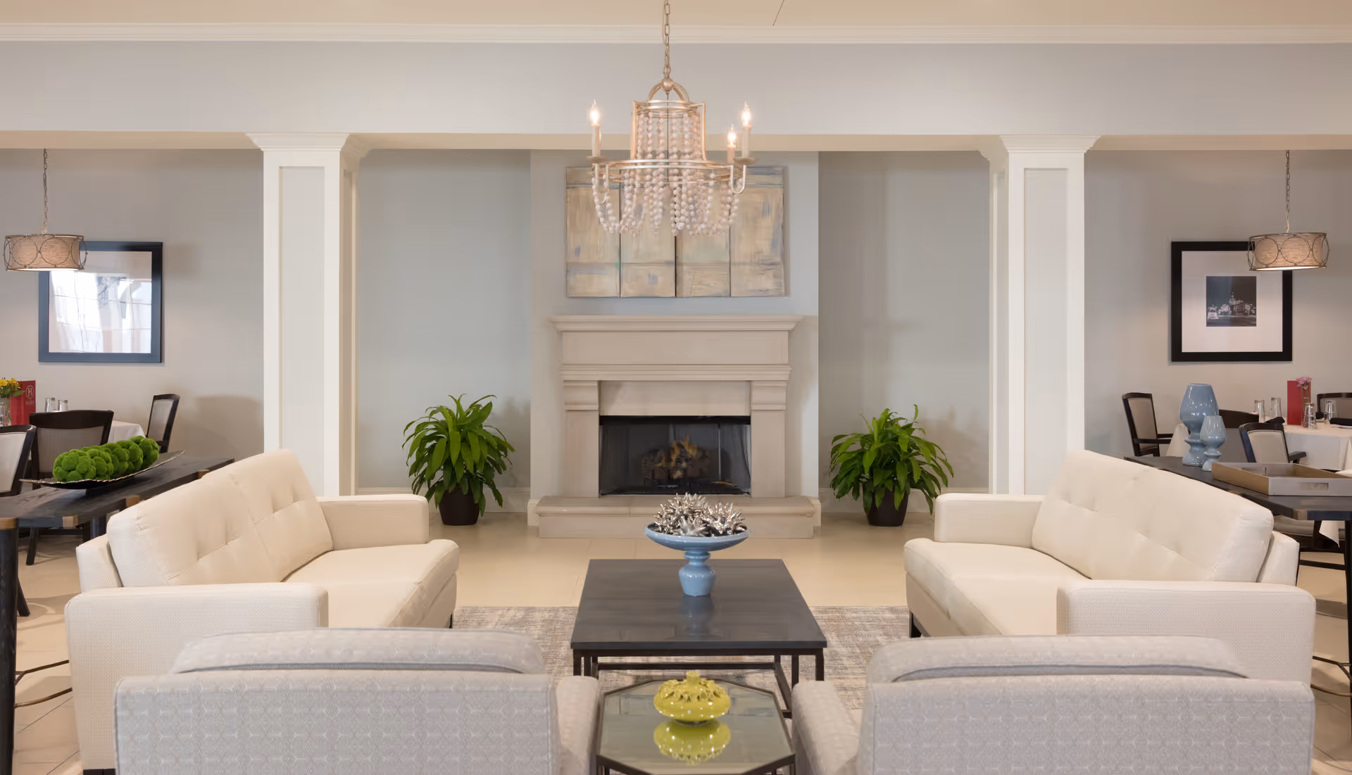 A bright and elegant living room area with two white sofas facing each other, a dark coffee table in the center with decorative items, a fireplace in the background flanked by two green plants, and a chandelier hanging from the ceiling. There are dining tables and chairs visible on both sides of the room with framed pictures on the walls.