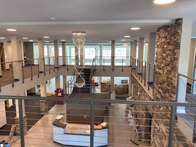 Interior view of a spacious, modern two-story common area with a large stone accent wall, metal railings, and a contemporary chandelier hanging from the ceiling. The area appears to be under construction or setup, with boxes and materials scattered around the floor and a reception desk in the center.