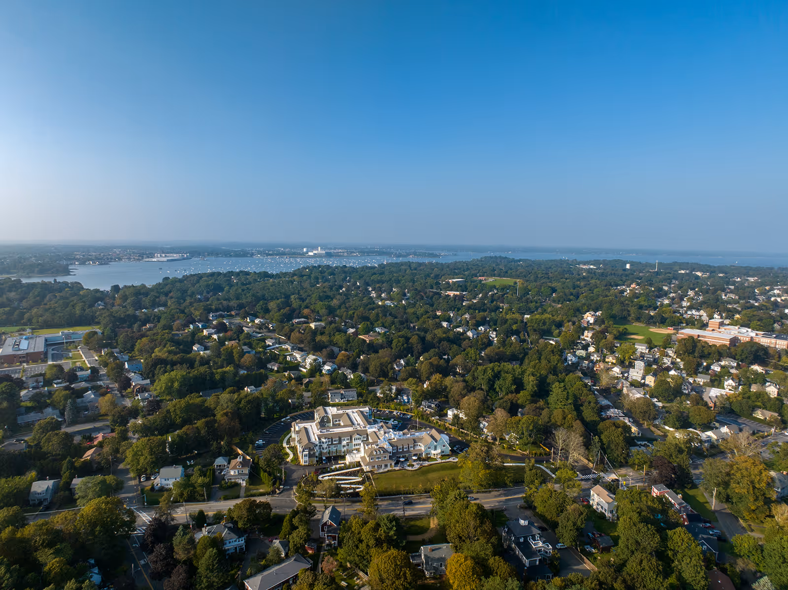 Aerial view of a suburban area with dense tree coverage and numerous houses. In the center foreground, there is a large building complex with a parking lot, likely The Mariner Marblehead facility. Beyond the residential area, there is a large body of water with boats visible under a clear blue sky.