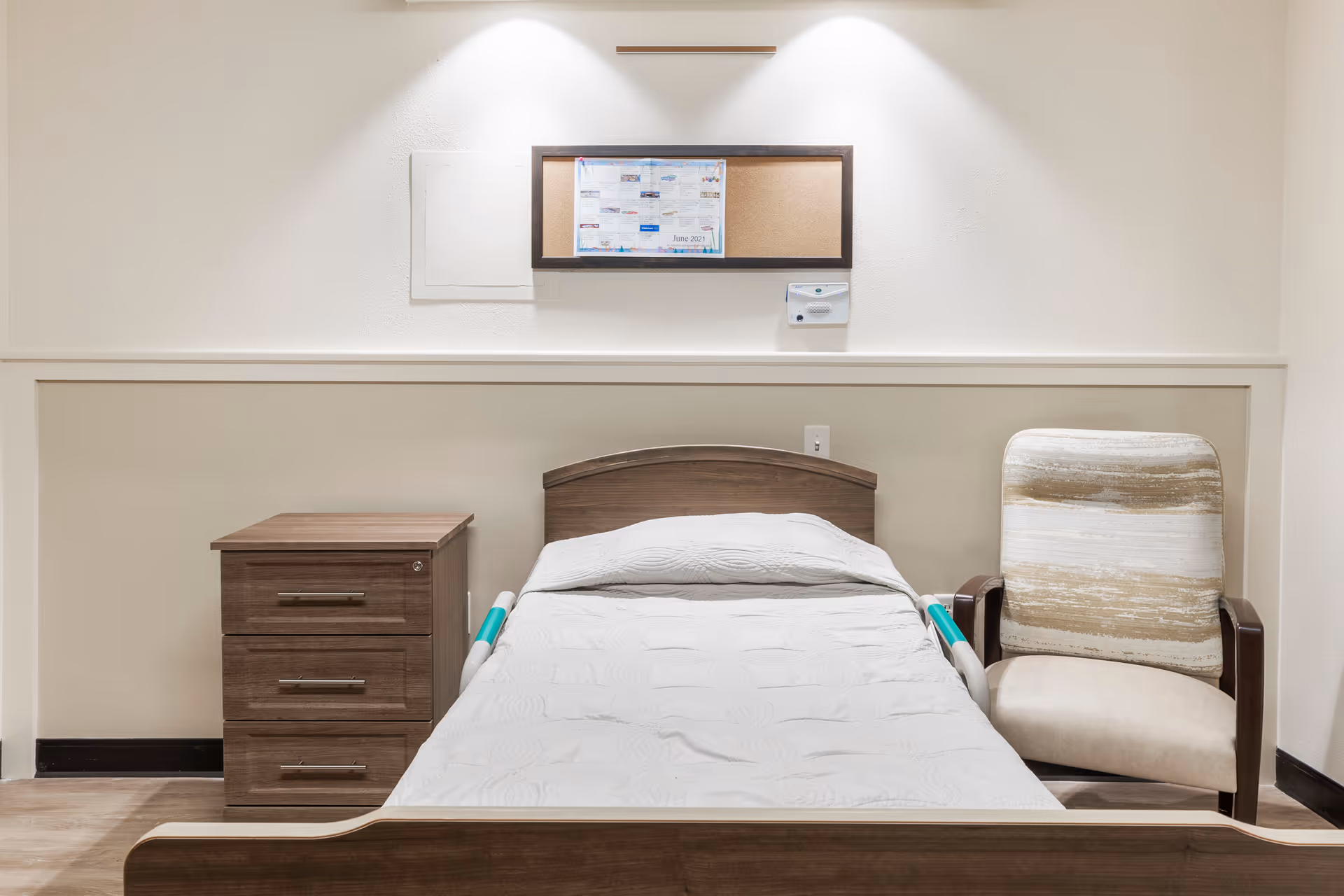 A simple bedroom in a senior living facility featuring a single bed with white bedding, a wooden nightstand with three drawers on the left, and a cushioned armchair with a striped pattern on the right. Above the bed is a bulletin board with a calendar and a wall-mounted device, all set against a beige and white wall.