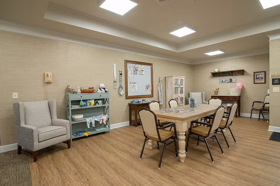 A well-lit room with a wooden table surrounded by six chairs, a gray armchair, a green shelf with decorative items, a whiteboard with drawings, and a small cabinet. The walls are beige with a wooden floor, and there is a sign that says 'LAUNDRY' on a shelf above a small table in the corner.