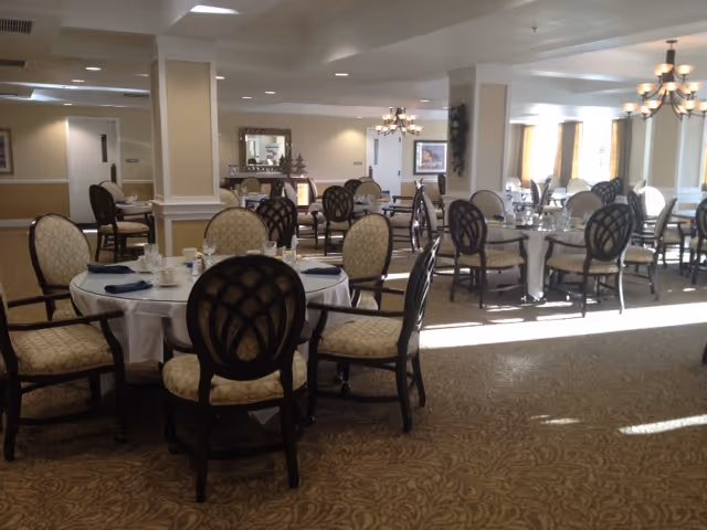A spacious dining room with multiple round tables covered with white tablecloths, each surrounded by cushioned chairs with patterned upholstery. The room is well-lit with natural light coming through large windows and elegant chandeliers hanging from the ceiling. The carpet has a subtle pattern, and the walls are painted in neutral tones with decorative molding.