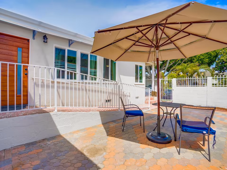 Sunlit outdoor patio with a large umbrella, table and chairs next to a white building entrance and ramp.