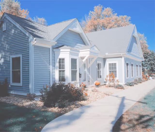 Exterior view of a single-story building with light blue siding and white trim, featuring multiple windows and a small covered entrance. The building is surrounded by landscaped bushes and a concrete walkway, with trees in the background under a clear blue sky.