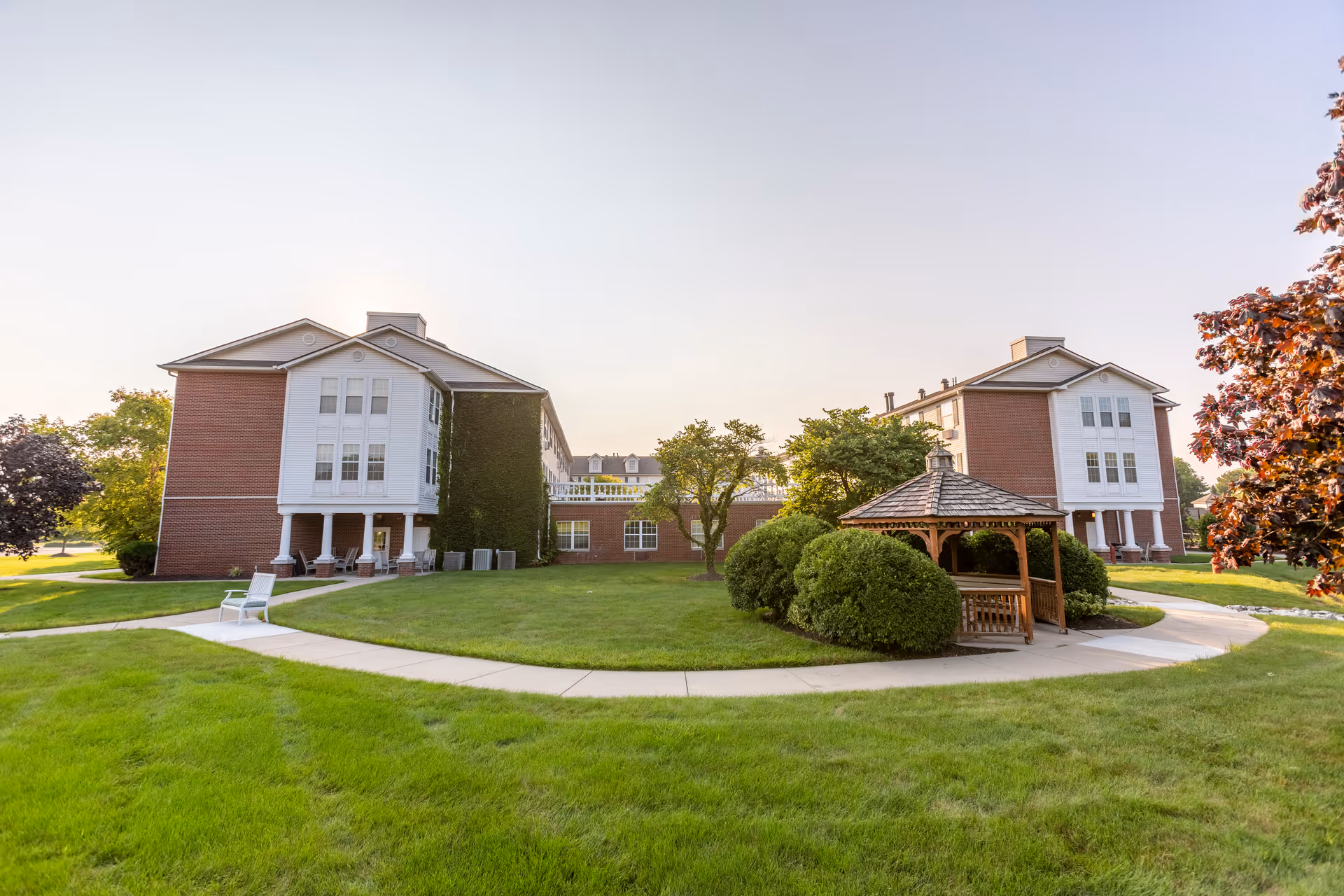 Outdoor view of Independence Village of Fishers South featuring two brick buildings with white accents, a well-maintained green lawn, a wooden gazebo surrounded by bushes, and a curved concrete walkway under a clear sky.