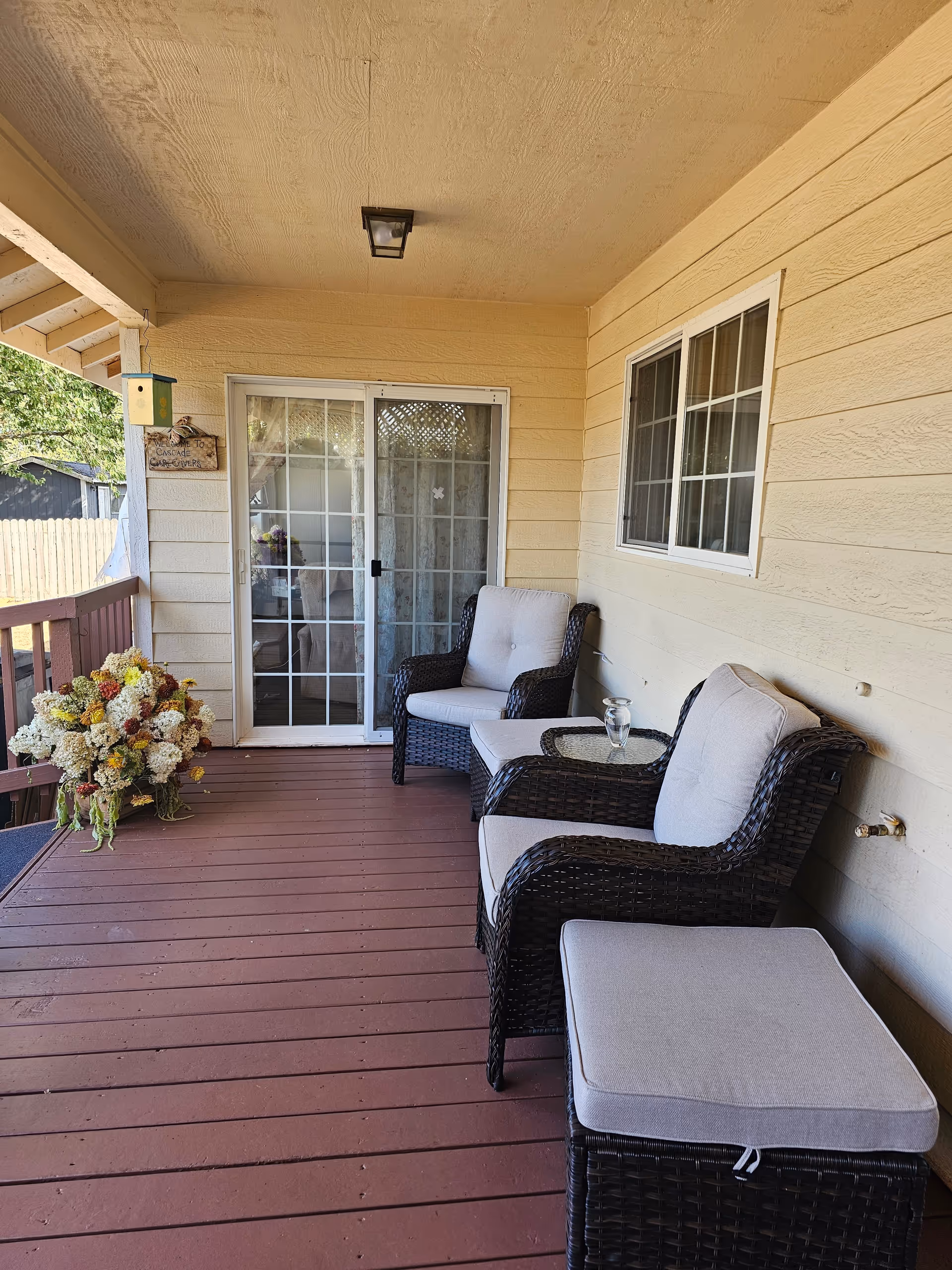 Covered outdoor porch area with two cushioned wicker chairs, a matching cushioned ottoman, a small glass-top side table with a glass of water, a sliding glass door, a window, and a large bouquet of flowers on the wooden floor.