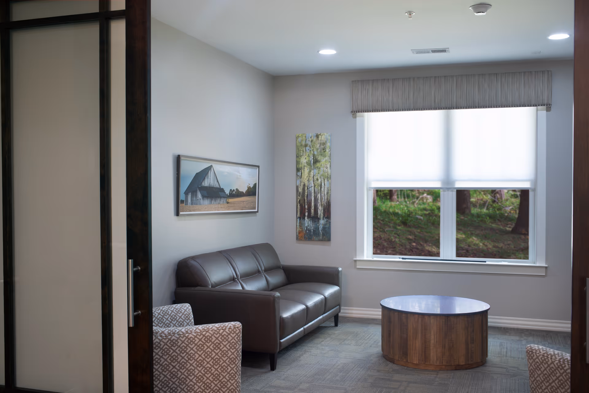 A small sitting area with a brown leather sofa, two patterned armchairs, and a round wooden coffee table. The room has light gray walls with two paintings, one of a barn and another of trees. A window with a white roller shade partially open reveals greenery outside.