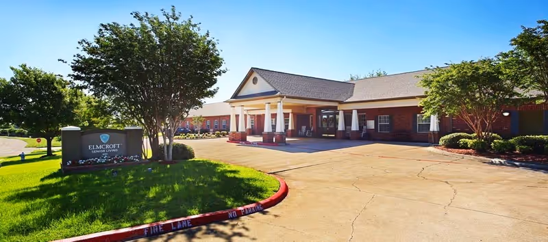 Exterior view of a senior living facility with a brick building, a covered entrance supported by white columns, and a driveway. There is a green lawn with trees and a sign that reads 'ELMCROFT SENIOR LIVING'. The sky is clear and blue.