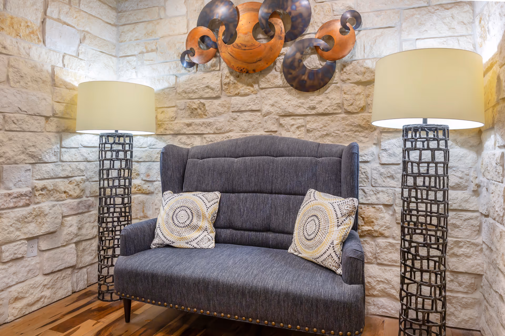 Cozy seating area with a gray loveseat, two patterned pillows, twin floor lamps, and a decorative stone wall with metal art.