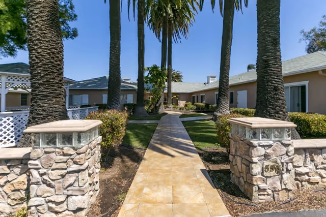 Tiled walkway lined with tall palm trees leading into a single-story courtyard with stone pillars and manicured landscaping.