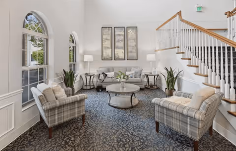 A bright and spacious living room area with two plaid armchairs, a gray sofa, and a round coffee table in the center. The room features large arched windows letting in natural light, two side tables with lamps, decorative plants, and three framed artworks on the wall. A staircase with wooden handrails and white balusters is visible on the right side.