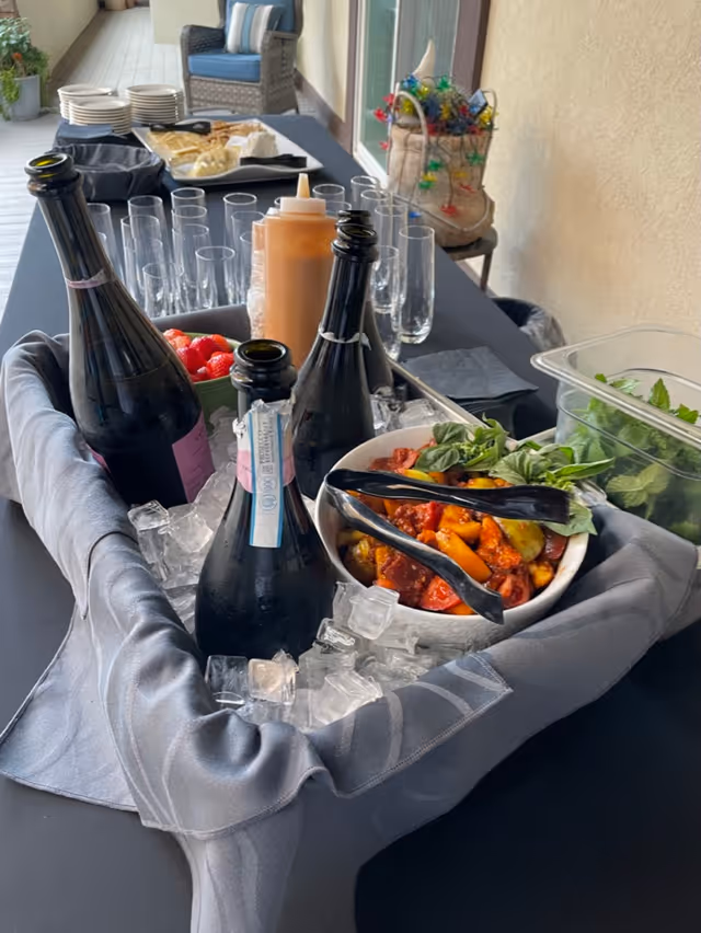 A table set up for a gathering with bottles of sparkling beverages chilling in a container filled with ice, a bowl of mixed tomatoes and basil with tongs, a container of fresh greens, and a tray with plates, cheese, and crackers in the background. There are also empty champagne flutes and a squeeze bottle of sauce on the table, which is covered with a black tablecloth. A wicker chair and a decorative item with colorful lights are visible in the background.