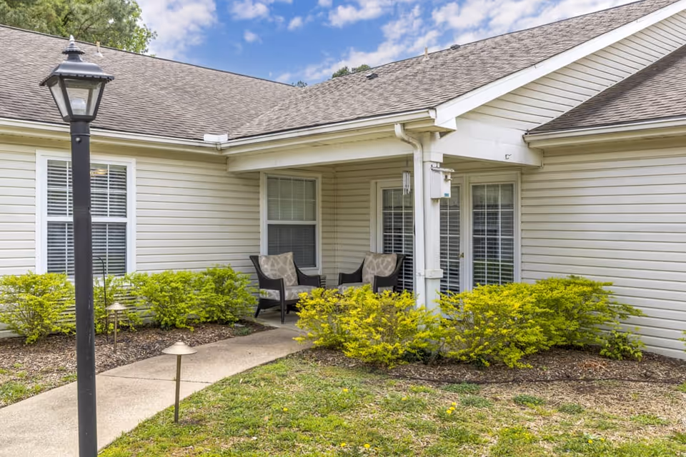 Covered patio with two chairs and a small walkway in front of a beige siding building surrounded by shrubs.