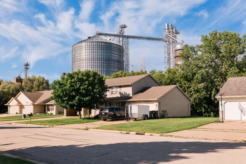 Suburban houses on a tree-lined street with a large industrial grain silo and metal structures visible behind them under a blue sky.