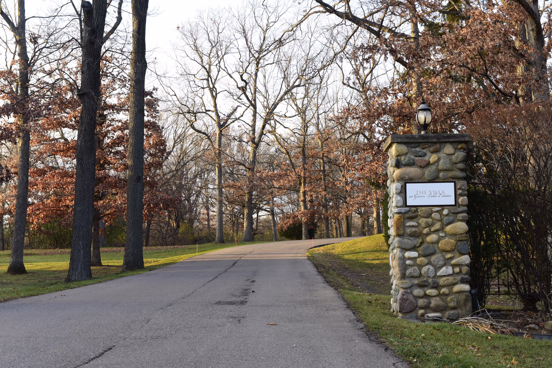 A paved driveway lined with trees on both sides leading into a wooded area. On the right side of the driveway, there is a stone pillar with a lamp on top and a sign that reads 'THE VILLA at Green Lake Estates'. The trees have brown leaves, indicating late autumn or early winter.