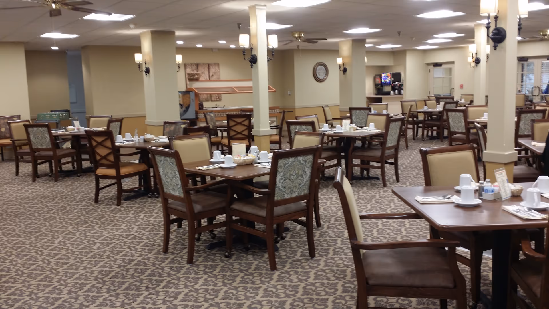 A spacious dining room with multiple wooden tables and chairs arranged neatly. Each table is set with white cups, plates, and napkins. The room has beige walls, patterned carpet flooring, and ceiling lights. There are columns and wall sconces providing additional lighting, and a beverage station is visible in the background.