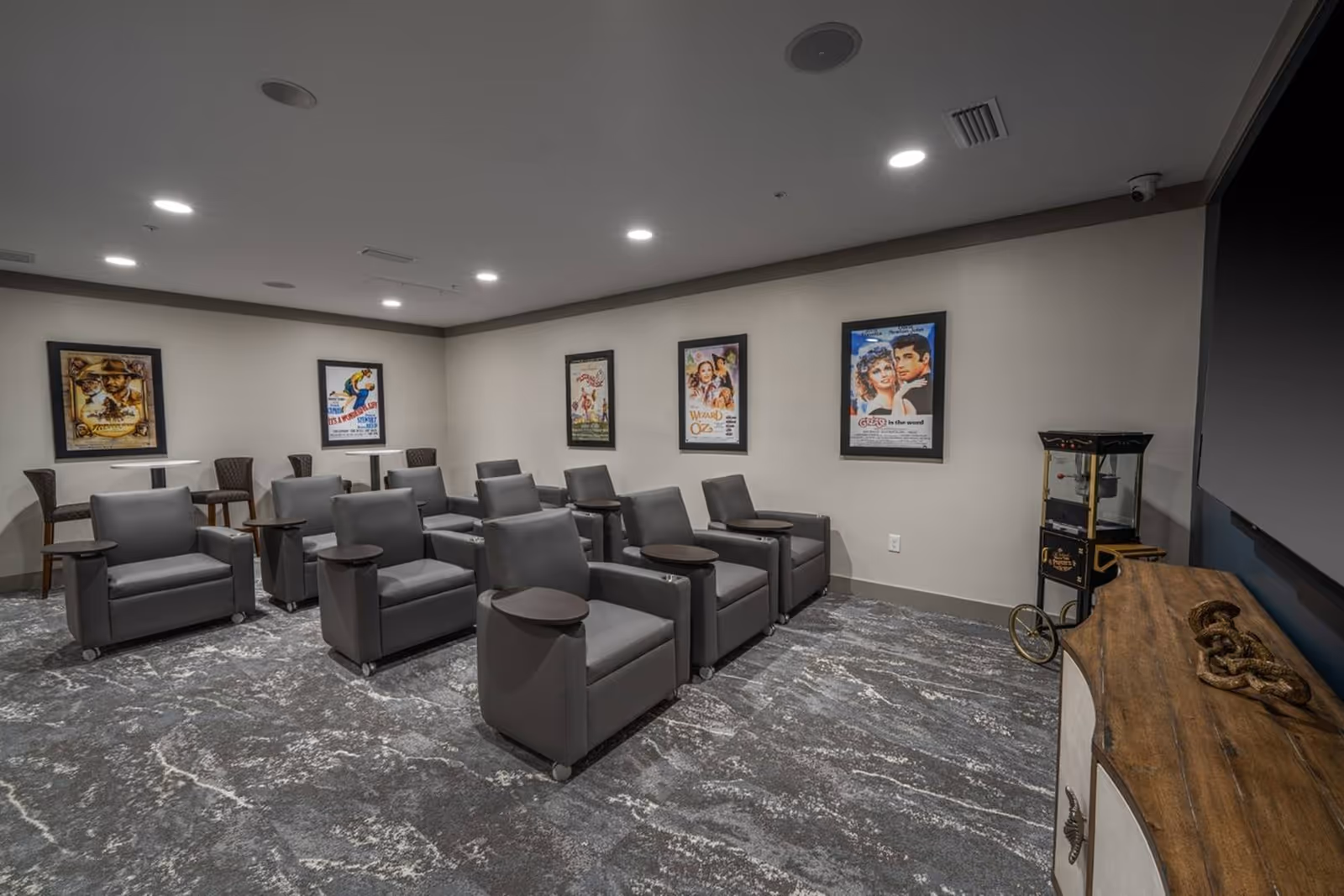 Small screening room with rows of gray recliner chairs, movie posters on the walls and a popcorn machine.