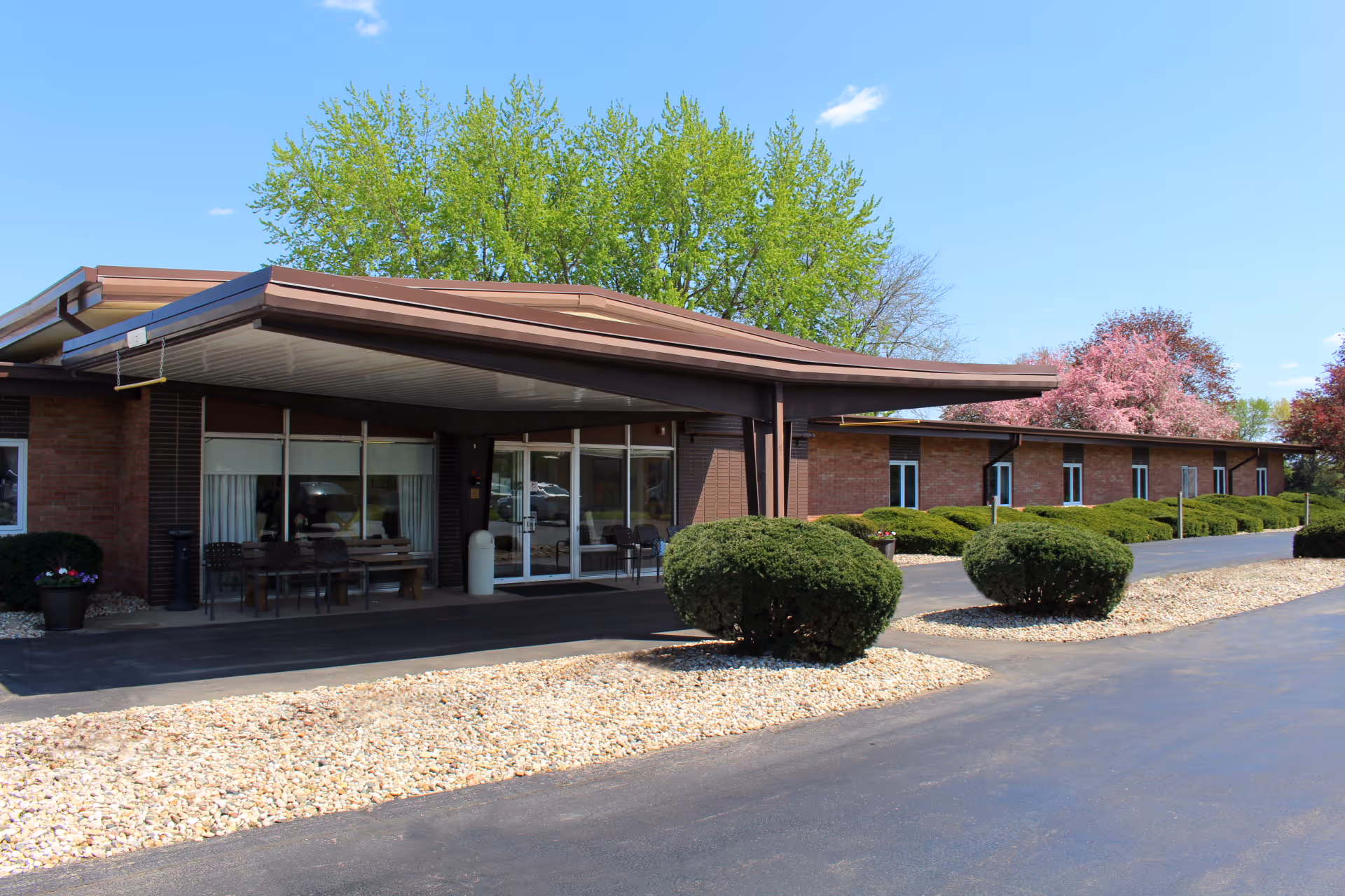 Exterior view of a single-story brick building with a covered entrance, surrounded by neatly trimmed bushes and trees with green and pink foliage under a clear blue sky.