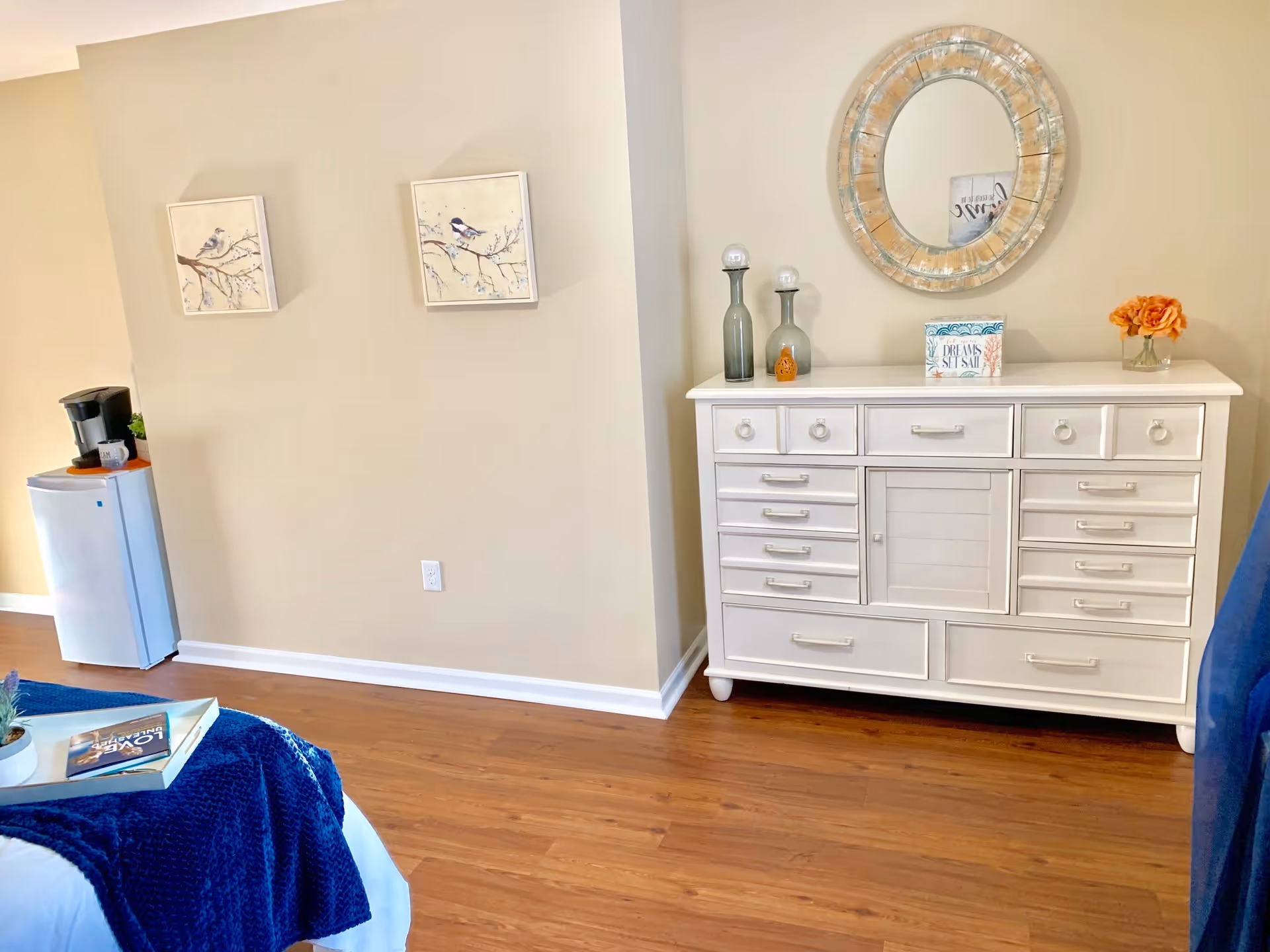 A cozy interior corner of a room featuring a white dresser with multiple drawers, decorated with two glass bottles, a small sign, and a vase with orange flowers. Above the dresser is a round mirror with a rustic frame. On the adjacent wall, there are two framed pictures of birds on branches. A small refrigerator with a coffee maker on top is positioned in the corner. The floor is wooden, and part of a bed with a blue blanket and a tray with a book and a small plant is visible in the foreground.
