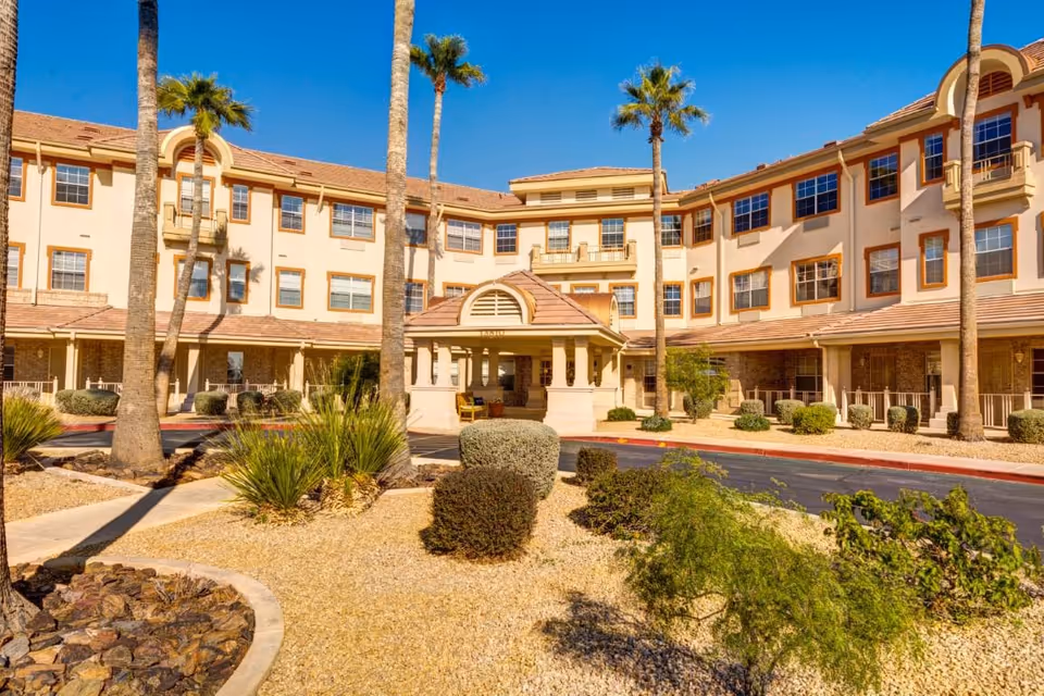 Exterior view of a multi-story assisted living facility with beige walls and brown trim, surrounded by desert landscaping including palm trees, bushes, and gravel. The building has multiple windows and a covered entrance with seating.