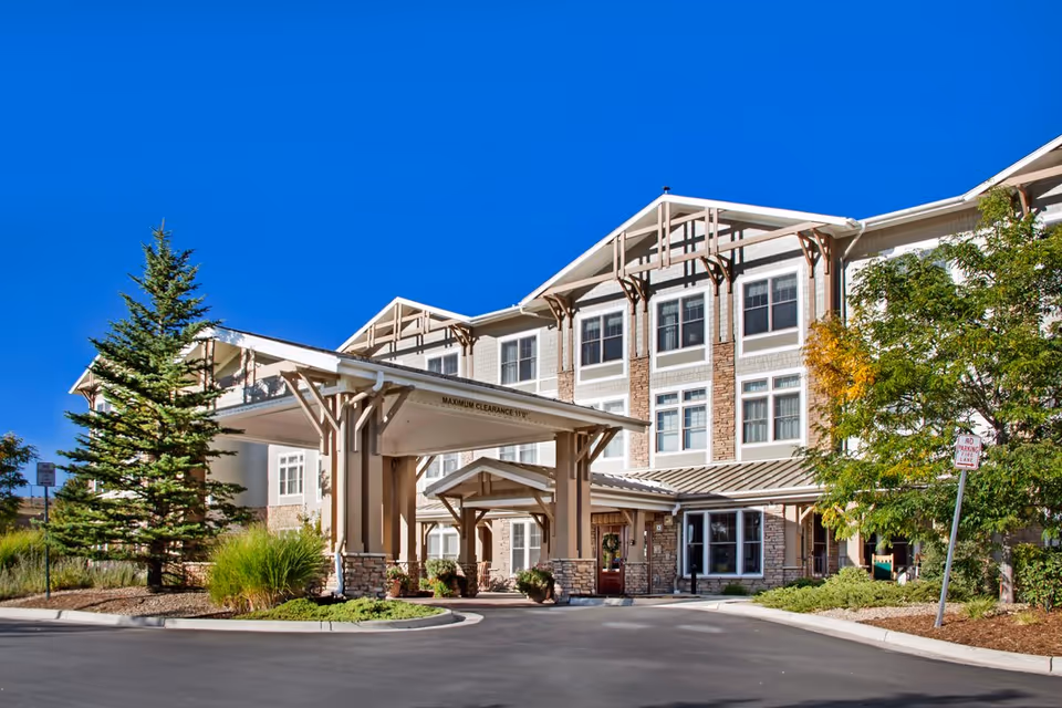 Exterior front view of a multi-story senior living facility building with a covered entrance, stone and siding facade, surrounded by trees and landscaping under a clear blue sky.