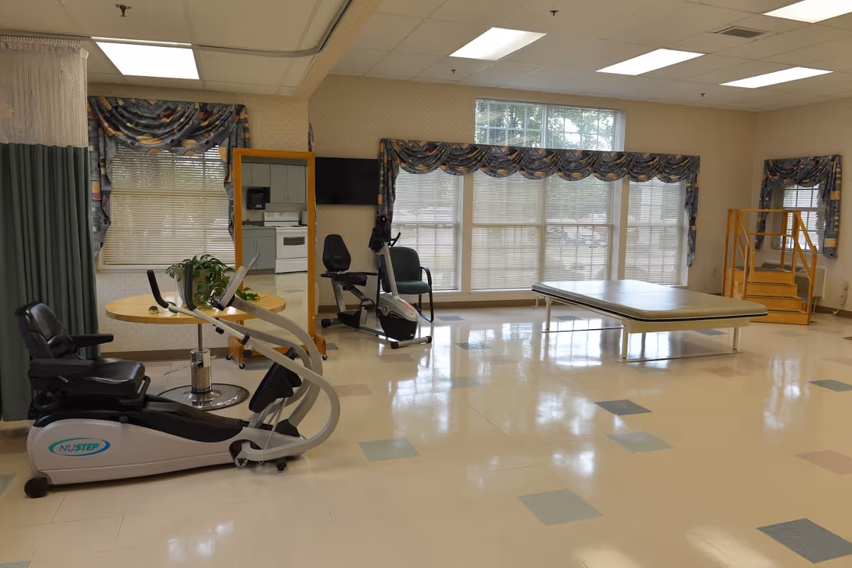A spacious room with exercise equipment including a NuStep recumbent cross trainer and a stationary bike, a padded therapy table, a small round table with a plant, and a set of wooden stairs with a handrail. The room has large windows with patterned valances, a wall-mounted TV, and a mirror reflecting a kitchen area.