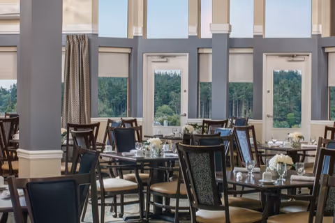 A bright dining room with multiple tables and chairs arranged neatly. Each table is set with white flowers in small vases, glassware, and white cups. Large windows and glass doors provide a view of green trees outside, allowing natural light to fill the room.