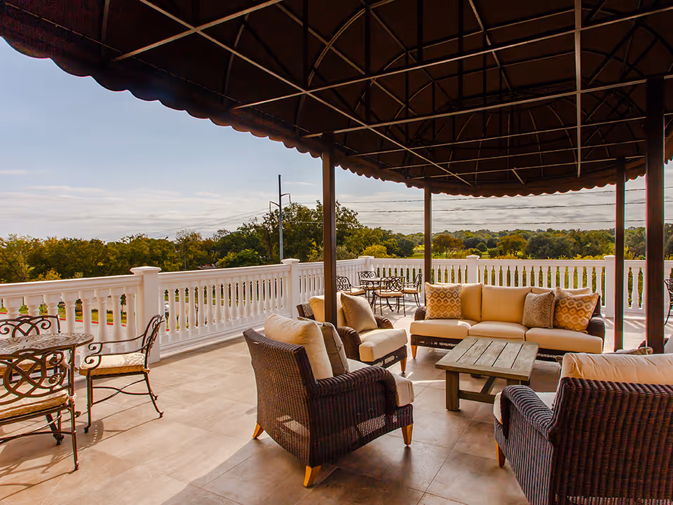 Outdoor patio area with cushioned wicker chairs, a sofa, a wooden coffee table, and metal dining tables with chairs under a large canopy, overlooking a scenic view of trees and open sky.