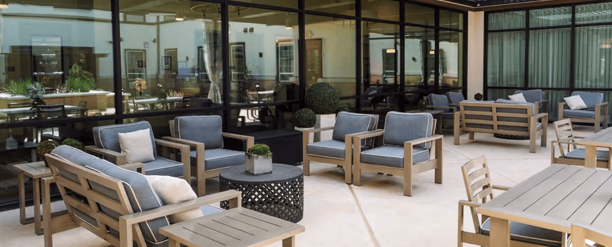 Outdoor patio area with modern wooden seating furniture including cushioned armchairs, sofas, and tables arranged on a concrete floor in front of large glass windows reflecting the interior of the building.
