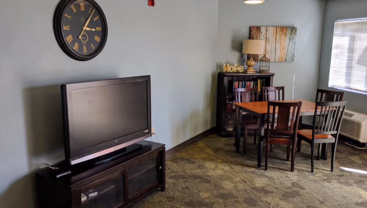 Small common room with a TV on a stand, a large wall clock, and a wooden dining table with chairs by a bookshelf and window.