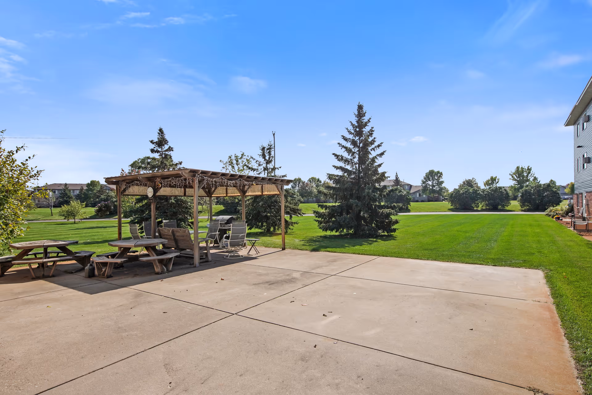 Outdoor patio area at Keller Lake Commons featuring a wooden pergola with seating underneath, picnic tables, and a large grassy lawn with trees in the background under a clear blue sky.